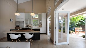 A modern kitchen with a central island, white chairs, and a dining table. There is a large window providing natural light and a view of the outdoors. - Your home away from home on the Farm, 4 Casitas Bed n Breakfast - Casita 1 (Cabo Rojo)