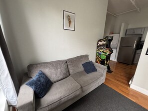 Een woonkamer met een grijze bank, twee blauwe kussens, een lijst met een foto en een kleurrijke spelcomputer.