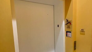 Un couloir étroit avec une porte d’ascenseur fermée, un détecteur d’incendie et un panneau indiquant le premier étage.