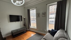 Un salon moderne doté d’un téléviseur à écran plat, d’un meuble de rangement sombre, d’un canapé gris, d’un lampadaire et de grandes fenêtres avec des rideaux.