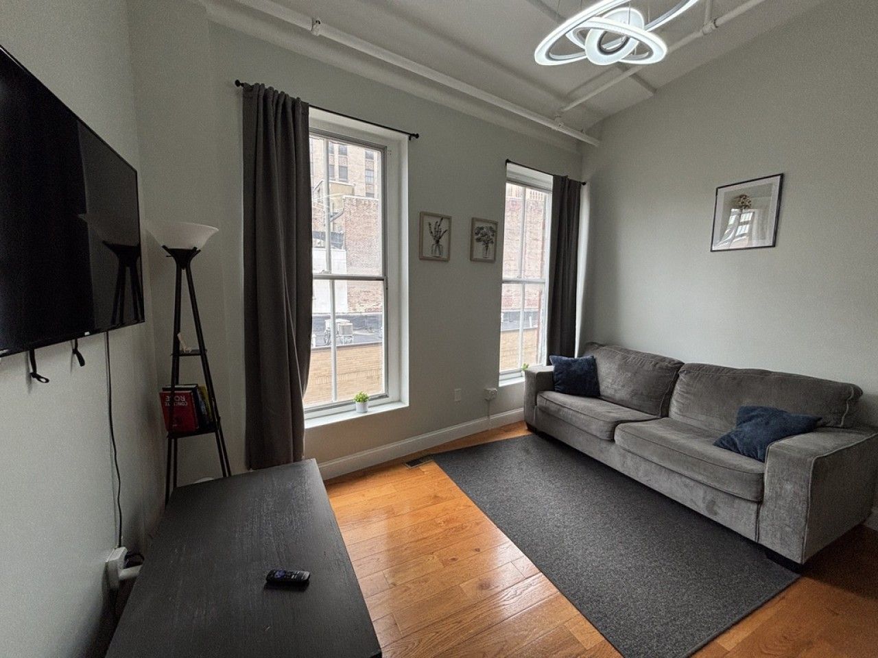 Un salon moderne avec un canapé gris, un téléviseur à écran plat fixé au mur et une grande fenêtre avec des rideaux.