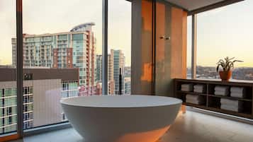 Un baño moderno con una bañera grande, un tocador de madera con toallas y vistas a la ciudad a través de amplios ventanales.