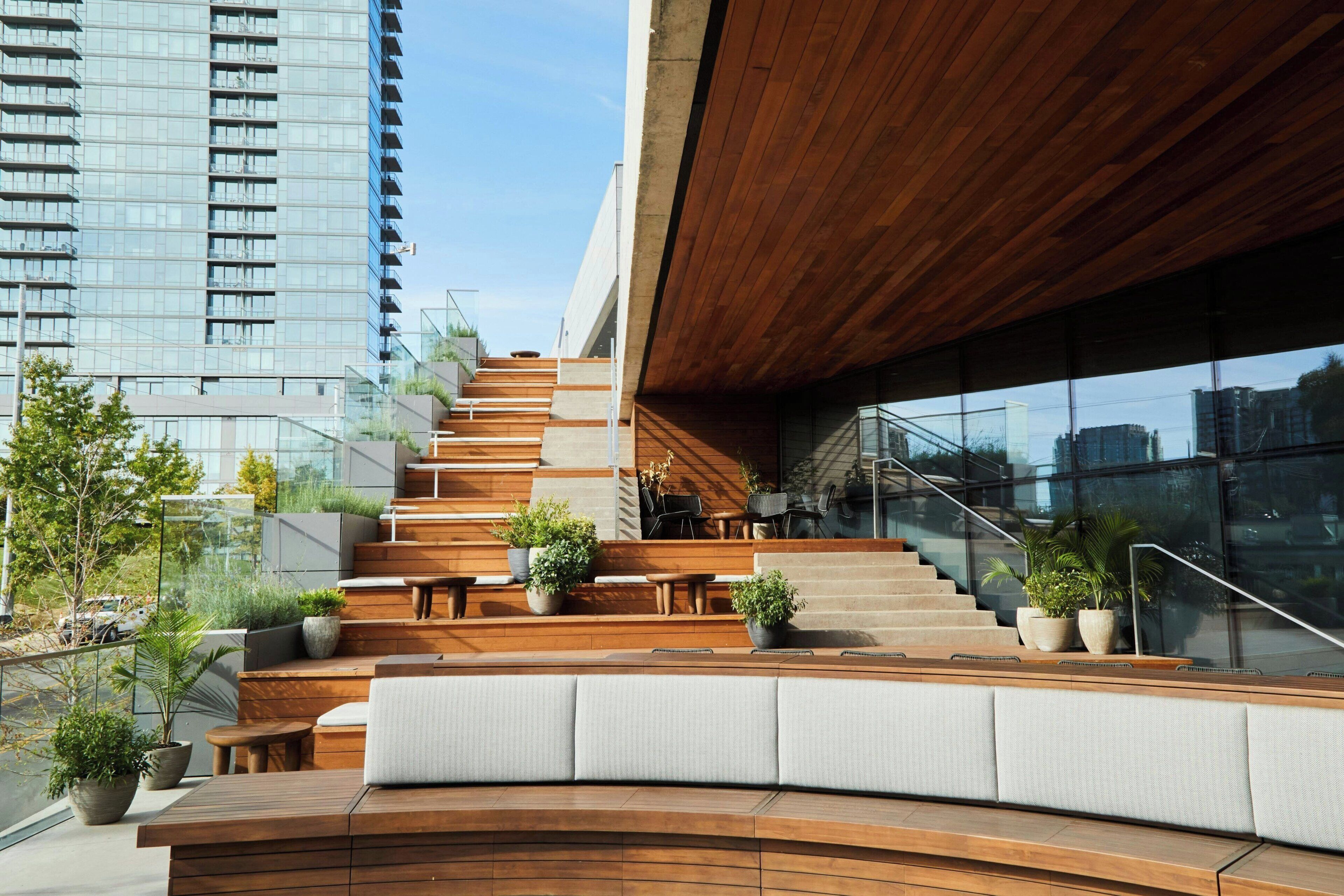 A modern outdoor seating area with wooden benches and planters, a staircase leading to an upper level, and a glass facade reflecting the cityscape.