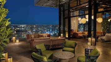 A rooftop lounge with a city view, furnished with sofas, chairs, and a round table.