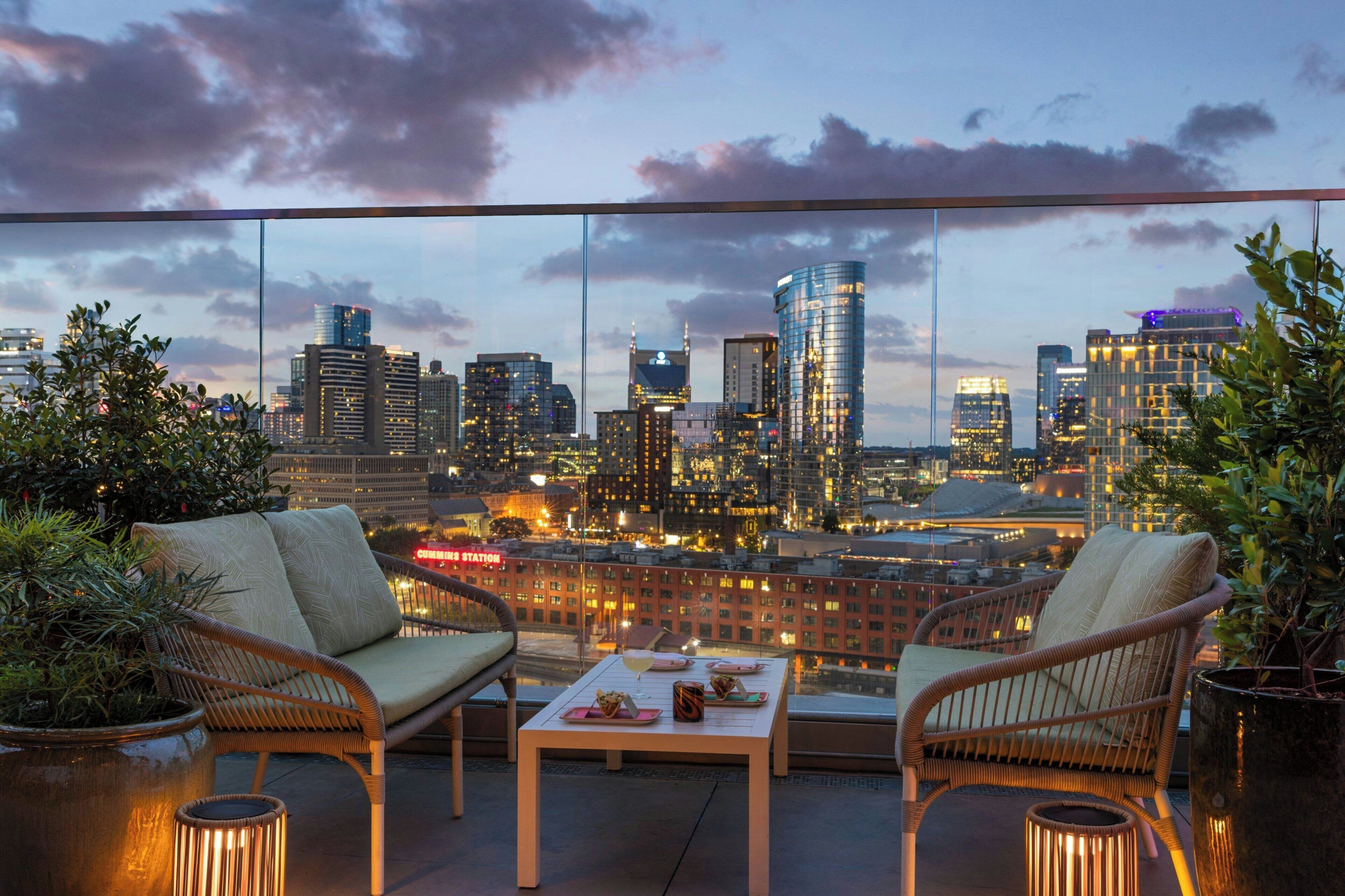 Terraza en la azotea con mobiliario de mimbre, una mesa con comida y vistas a un horizonte urbano.