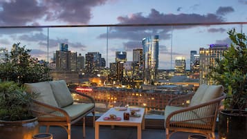 Terraza en la azotea con mobiliario de mimbre, una mesa con comida y vistas a un horizonte urbano.
