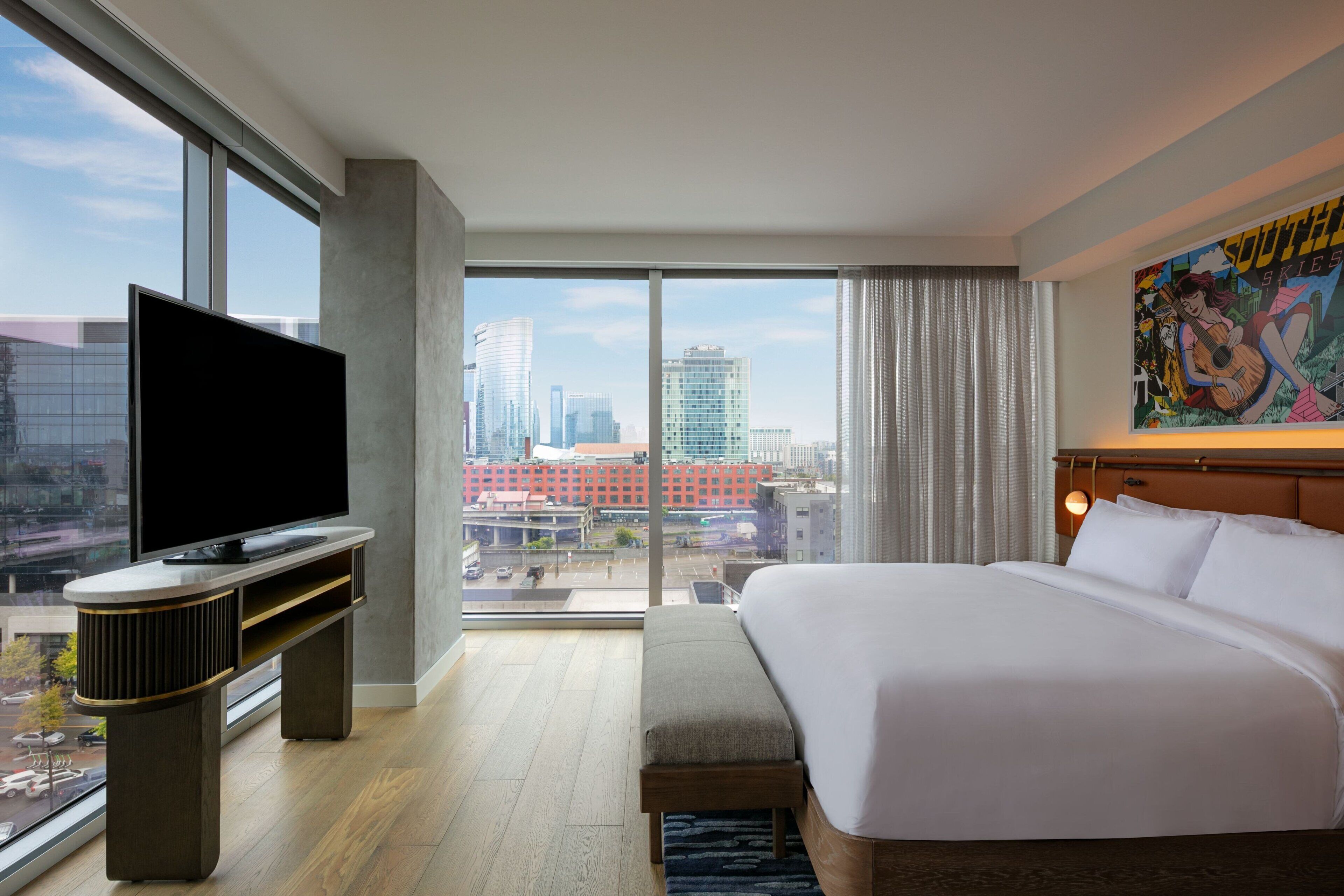 A hotel room with a large bed, a flat-screen TV, a city view, and a painting on the wall.