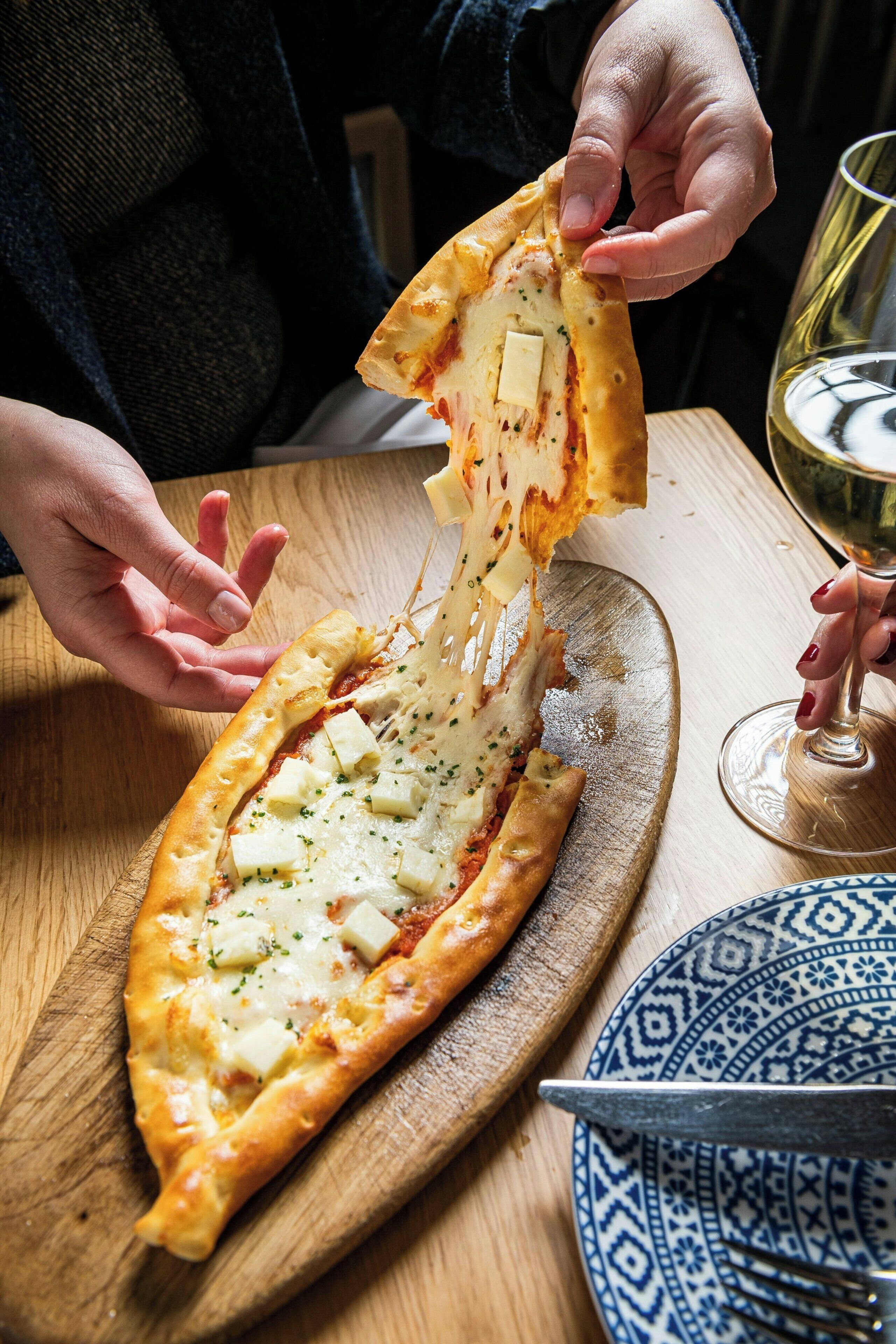 Une personne tenant une part de pizza garnie de fromage et d’herbes, un verre de vin et un couteau, le tout posé sur une table en bois.
