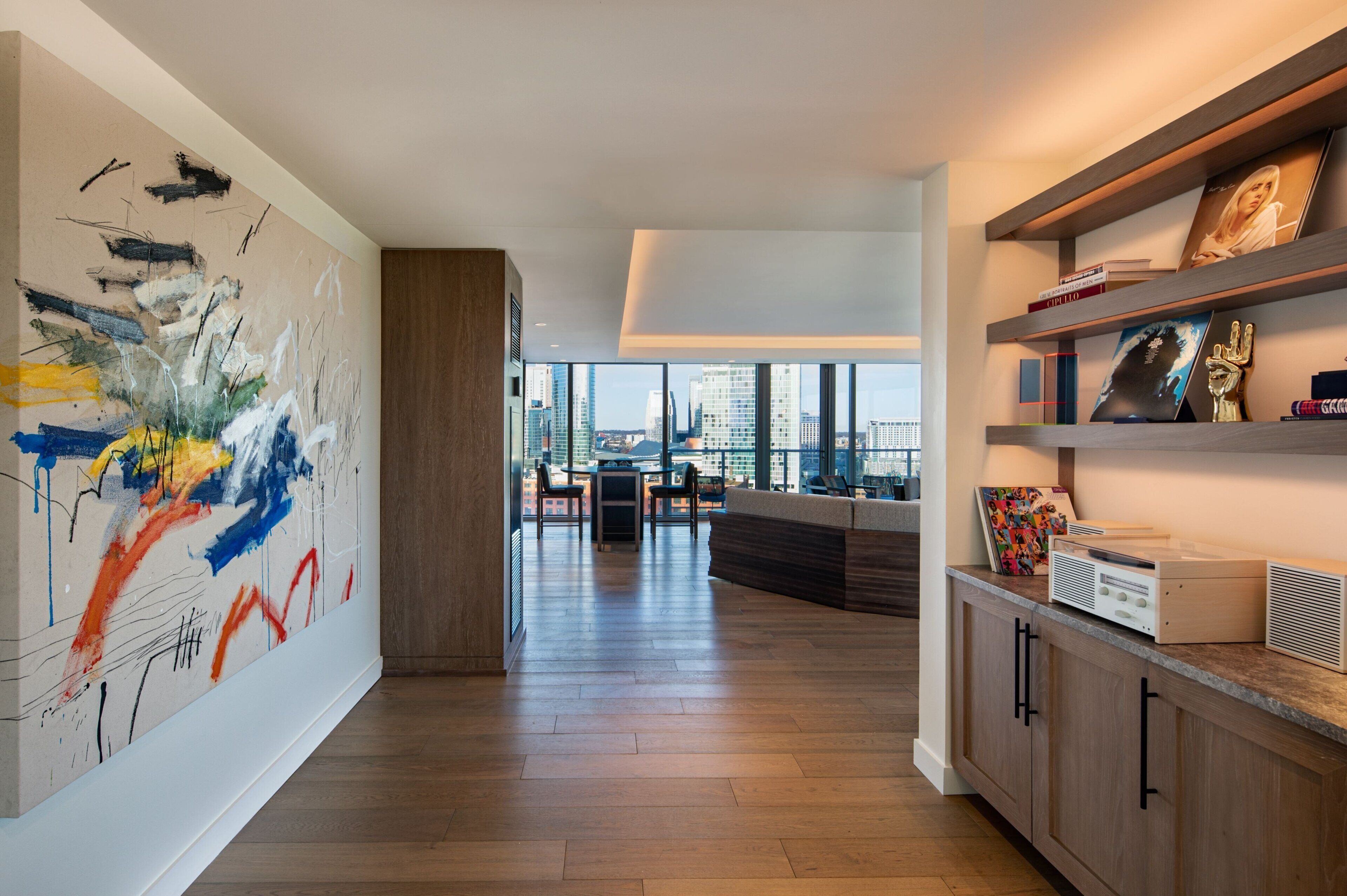 A modern interior with a large abstract painting, a wooden cabinet with a radio, and a city view through large windows.