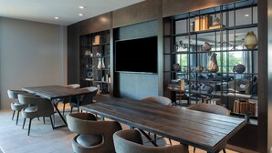 Un comedor moderno con una larga mesa de madera y varias sillas, un televisor de pantalla plana en la pared, y una estantería llena de objetos decorativos.