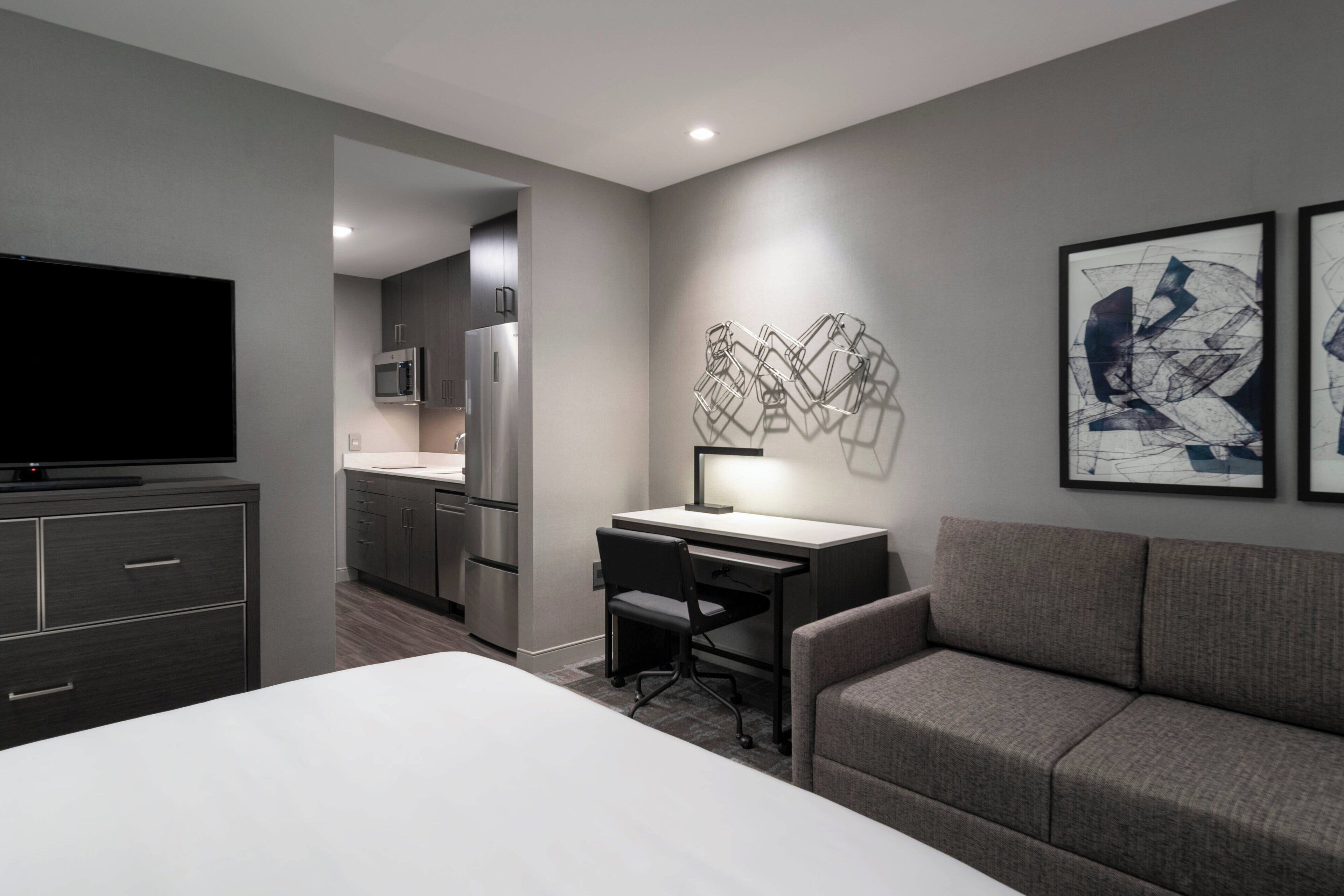 A modern hotel room with a large bed, a desk with a chair, a sofa, a flat-screen TV, a microwave, and a refrigerator.