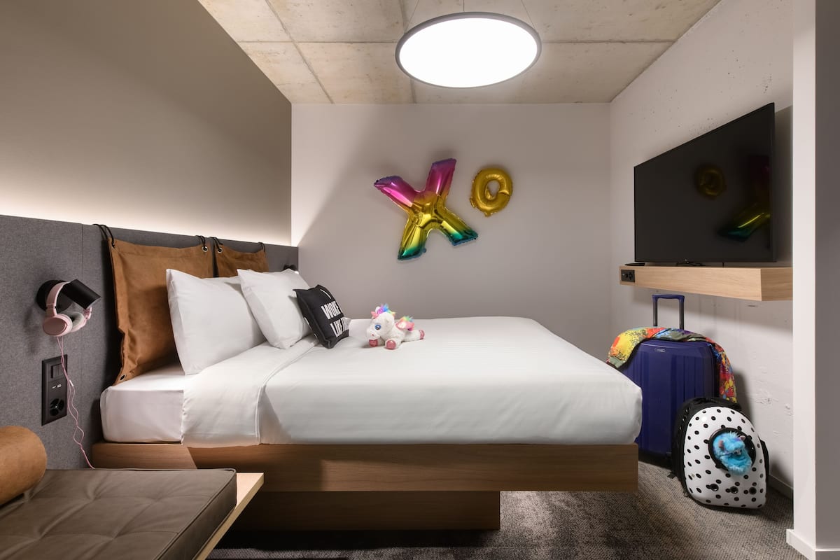 Quarto de hotel com cama, almofadas, um brinquedo de pelúcia, mala e balões.