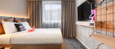 A modern hotel room with a large bed, a TV mounted on a wall, a neon flamingo sign, and a window with curtains.