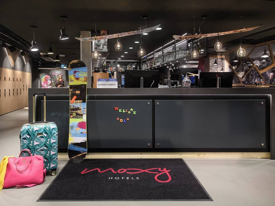 A modern hotel reception area with a black reception desk, a digital screen, and a welcome sign. There are suitcases and a pink handbag on the floor.