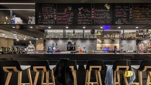 Zona de bar con una pizarra que muestra un menú de bebidas y sus precios. Hay taburetes de bar y una barra con una caja registradora.