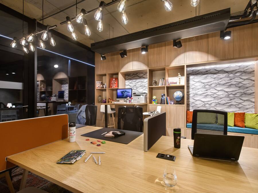 A modern office space with a wooden desk, a laptop, and various office supplies. There are cubicles, a sofa with cushions, and a decorative wall with a textured pattern.