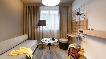 Ein modernes Hotelzimmer mit Sofa, kleinem runden Tisch, Lampe, Koffer und einer Wandablage.