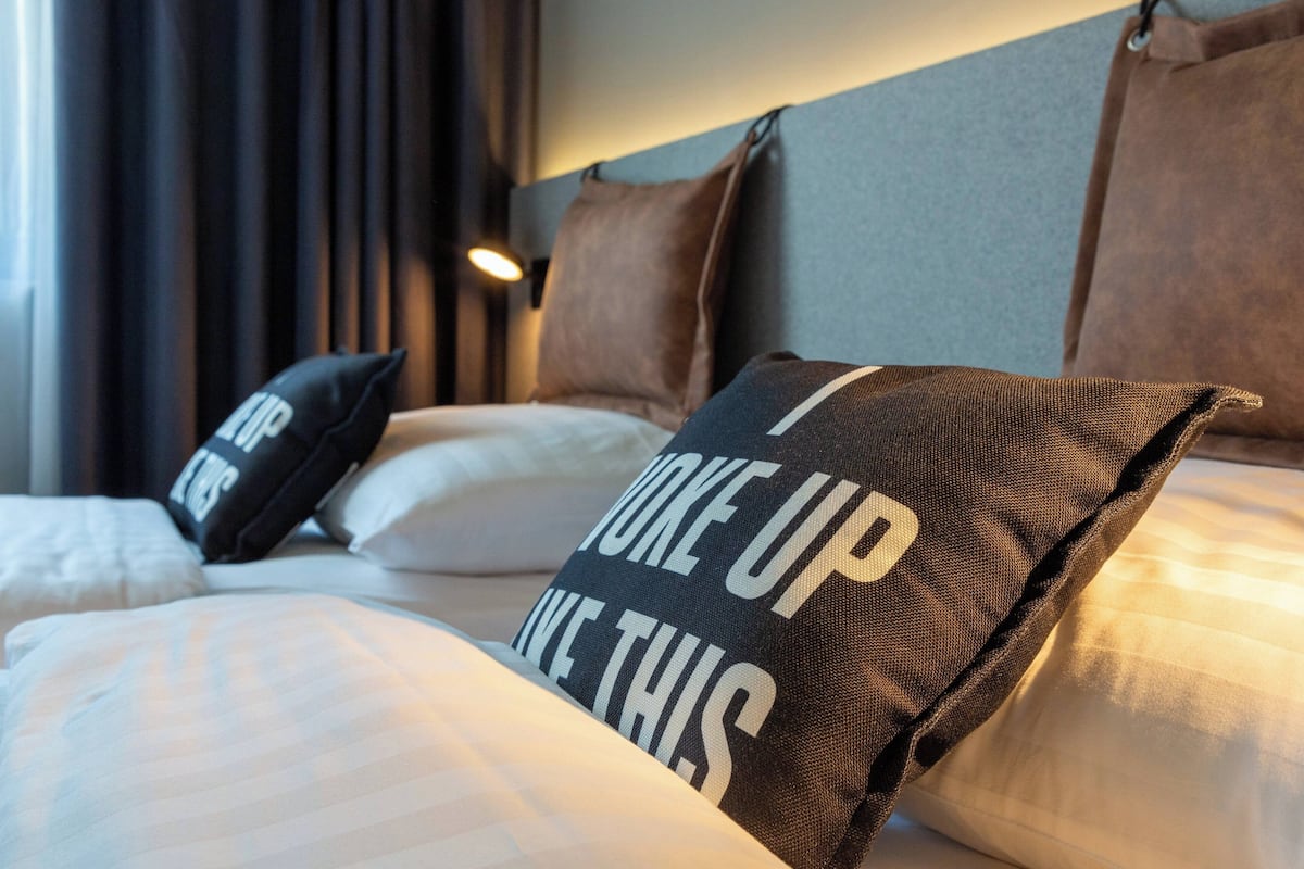 Quarto de hotel com cama, travesseiros com mensagens motivacionais, cabeceira e janela com cortinas.