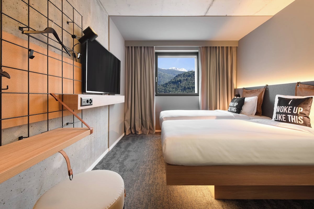 Quarto de hotel com duas camas, televisão numa parede, janela com vista para as montanhas e uma estante de madeira com uma TV.