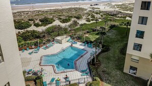Ein Poolbereich mit Blick auf Strand und Meer, umgeben von Grünpflanzen und einem Gebäude.