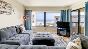 Ein modernes Wohnzimmer mit einem grauen Sofa, einem schwarzen Ottomanen und einem Flachbildfernseher. Ein großes Fenster bietet Blick auf den Strand und die Gebäude.