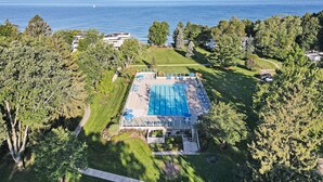 An aerial view of a property with a swimming pool surrounded by trees, a clear view of the sea, and a well-maintained lawn.