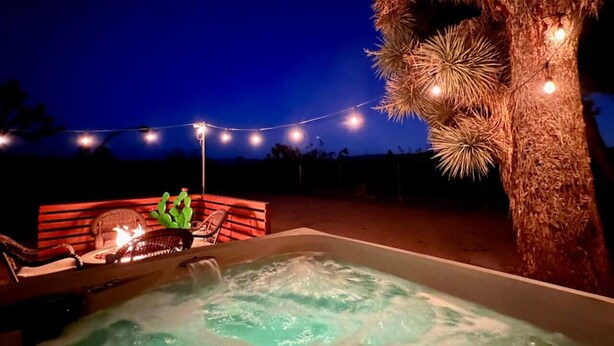 A hot tub with a wooden deck and chairs, surrounded by a string of lights and a large tree. - The Desert Peacock, Hot Tub, Central AC & VIEWS (Yucca Valley)