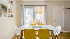 A dining area with a white table and yellow chairs, a window with curtains, and a door with a sign. - The Desert Peacock, Hot Tub, Central AC & VIEWS (Yucca Valley)