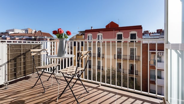 En balkon med et bord og stole, en vase med blomster og udsigt til bygninger.
