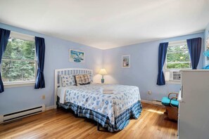 A bedroom with a bed, a nightstand, a lamp, a window with curtains, and a wall-mounted air conditioner. - Easy Bike Path Access, Dog Welcome! (Chatham)