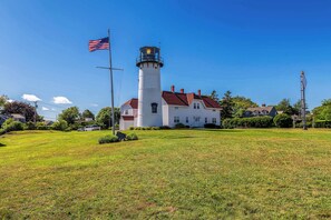 A lighthouse with a white exterior and red roof, surrounded by a grassy field and residential houses.