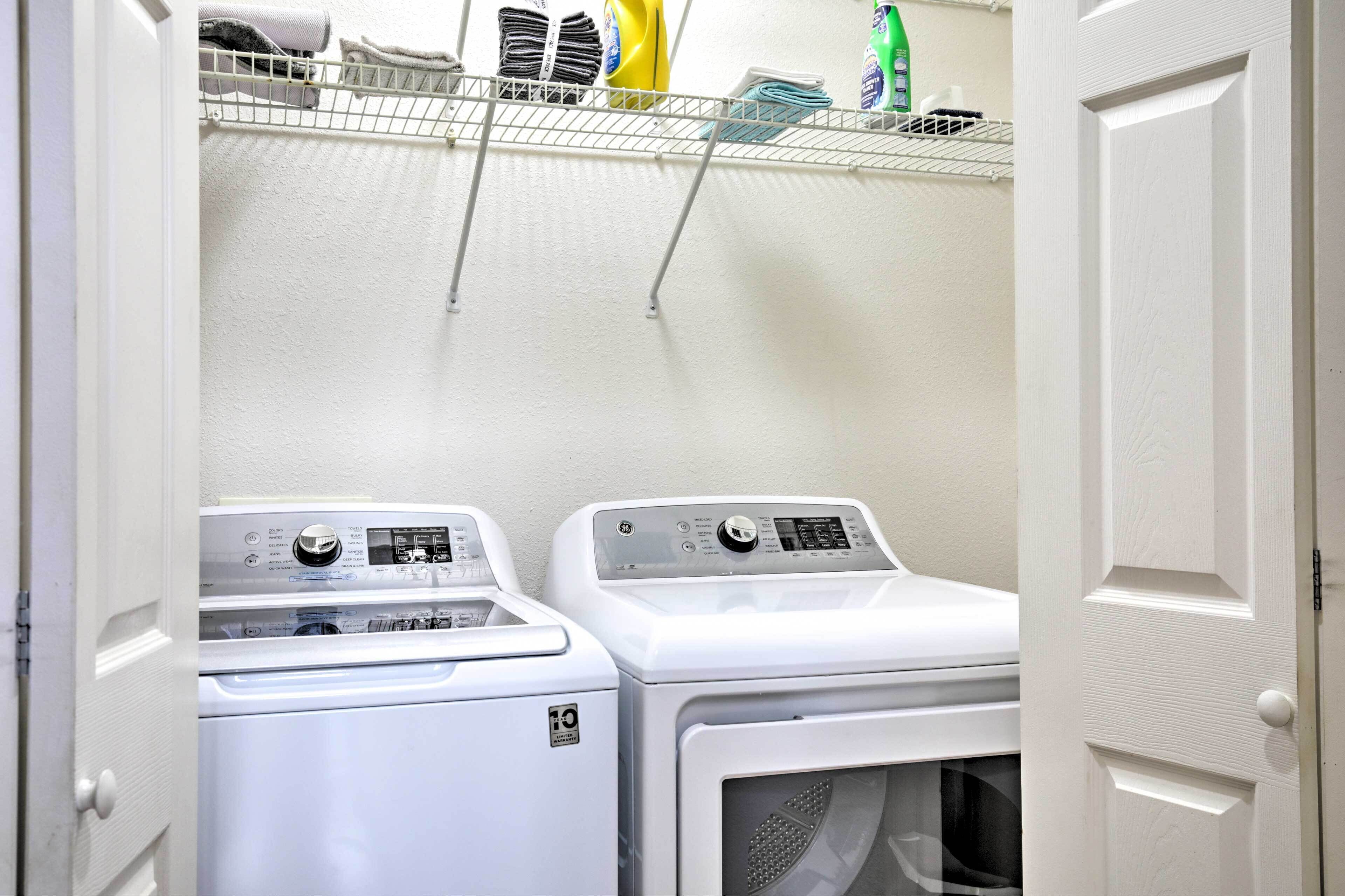 Une buanderie équipée de deux machines à laver et deux sèche-linge superposés, d’un étendoir métallique et d’étagères contenant des produits de lessive.