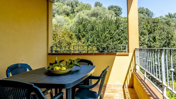 Un balcón con mesa y sillas, un cuenco de fruta y vistas a una ladera boscosa.