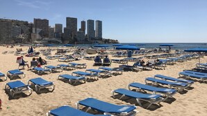 Ein Strand mit Reihen blauer Liegestühle und Sonnenschirme, im Hintergrund ein Stadtpanorama und ein klarer, blauer Himmel.
