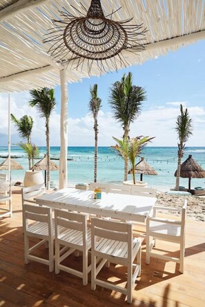 Ein Essbereich am Strand mit Tisch und Stühlen unter einem Reetdach mit Blick auf den Ozean.