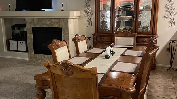 Comedor con una gran mesa de madera, seis sillas, un televisor sobre una chimenea y un aparador decorativo con puertas de vidrio.