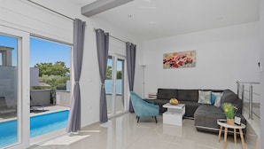 Un salon moderne avec une piscine, des rideaux gris, un canapé gris, un fauteuil turquoise et une petite table sur laquelle est posée une plante.