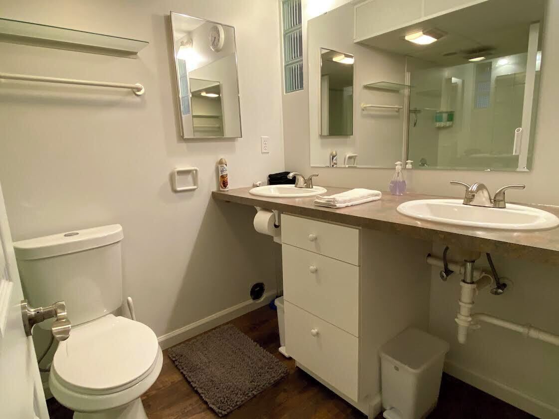 Une salle de bain avec des toilettes, deux lavabos, un miroir et une petite poubelle.