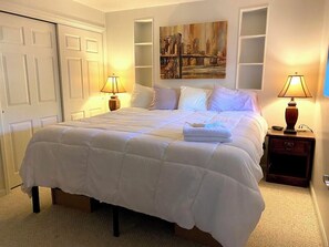A neatly made bed with white bedding, two bedside lamps, a painting above the bed, and a closed door to the left.
