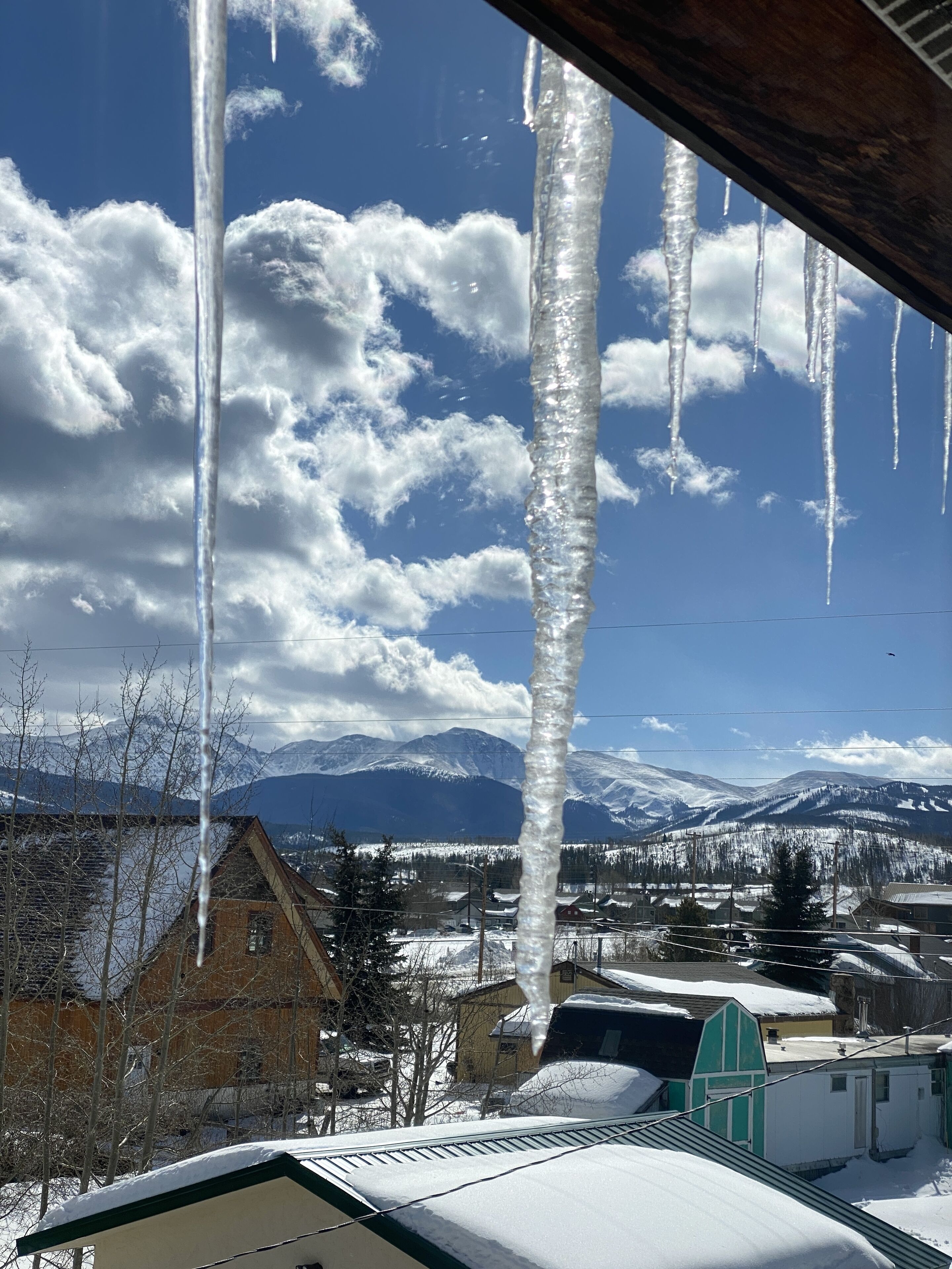 A snowy landscape with icicles hanging from a roof, mountains in the background, and a clear blue sky.