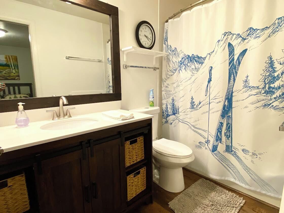 A bathroom with a white sink, dark wooden cabinets, a white toilet, a clock on the wall, and a shower curtain with a mountain design.