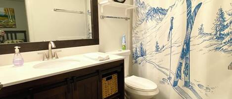 A bathroom with a white sink, dark wooden cabinets, a white toilet, a clock on the wall, and a shower curtain with a mountain design.
