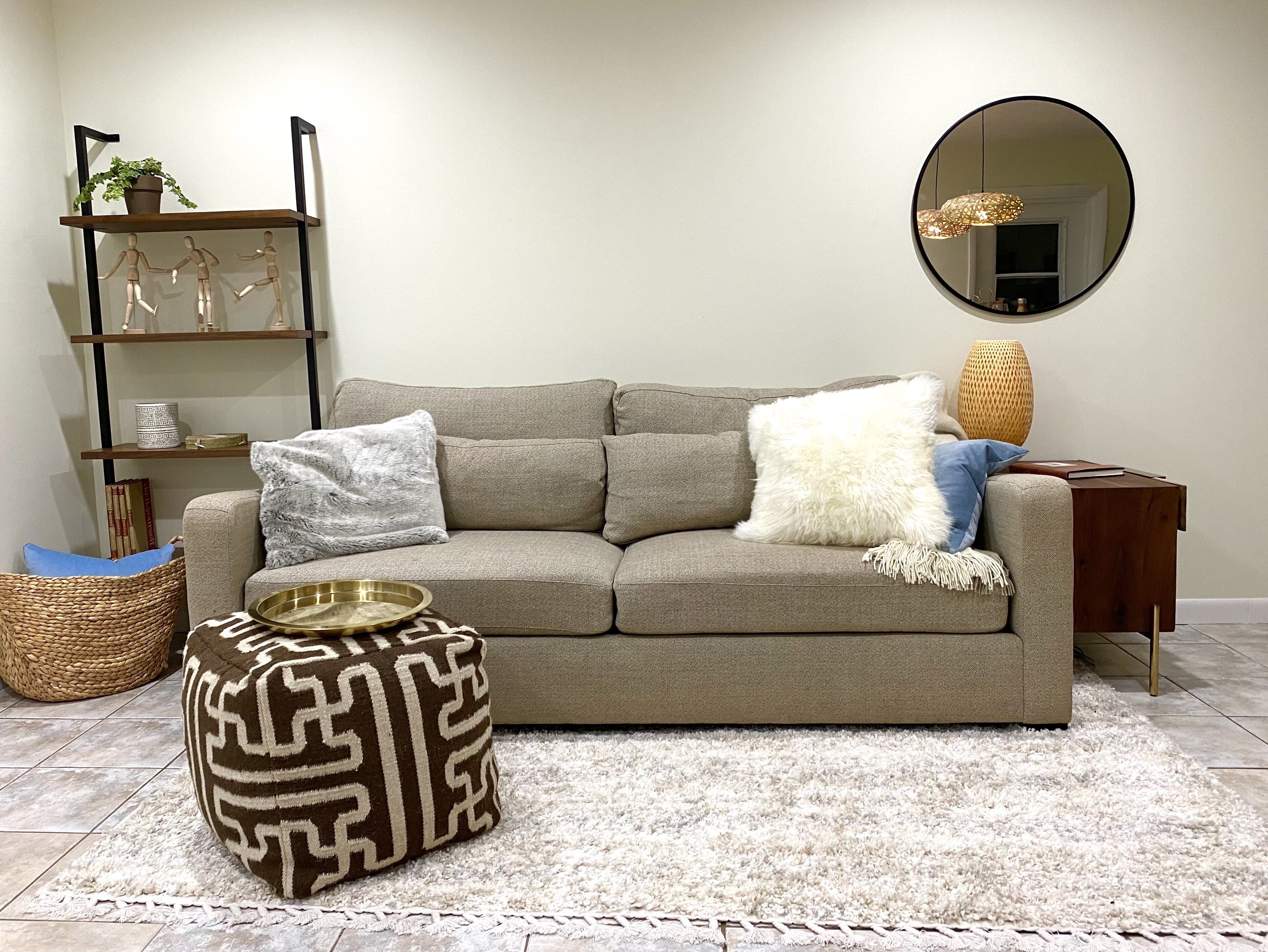 A neutral-toned living room with a sofa, a round mirror, a side table, and a woven pouf.