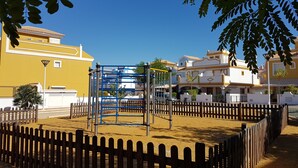 Un parque infantil con una estructura azul para escalar, rodeado por una cerca de madera, en una zona residencial con casas amarillas y blancas.