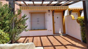 Un espacio al aire libre cubierto con una pérgola de madera, una puerta blanca y una ventana con rejas de metal.