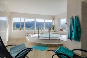 A spa room with a hot tub, two chairs, a window with a view of the forest, and a towel rack.