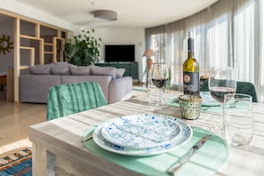 Una mesa de comedor dispuesta con platos, vasos y una botella de vino en un moderno salón con sofá y televisor.
