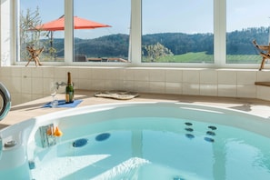 A hot tub with a view of a forested landscape through large windows.