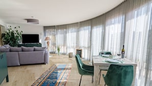 A modern living room with a sofa, a dining table with chairs, a TV, and a large window with sheer curtains.