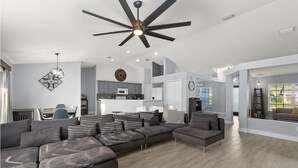 A spacious living area with a large sectional sofa, a ceiling fan, and a kitchen in the background. - St Lucie Central Cozy Casa (Port St. Lucie)